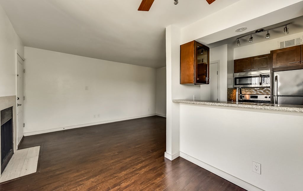 A modern apartment interior from our portfolio at Orand Capital features an open floor plan. The living area has a hardwood floor and a fireplace. The adjacent kitchen includes stainless steel appliances, dark wood cabinets, and a granite countertop bar. The walls are painted white.