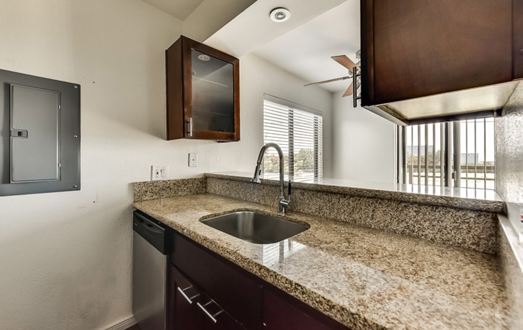 A modern kitchen from our portfolio features a granite countertop, stainless steel sink with a tall faucet, dark wooden cabinets, and a small window overlooking the room. A control panel is mounted on the white wall to the left, and vertical blinds cover sliding glass doors in the background.