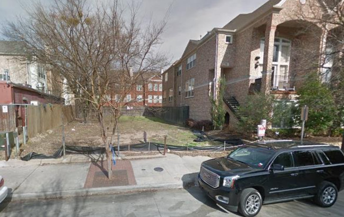 A vacant lot is seen between two brick buildings. The lot, part of Our Portfolio - Orand Capital, is surrounded by a temporary black fence and contains some bare trees and undergrowth. A black SUV is parked on the street in front of the lot. The sky is cloudy, and the scene appears quiet.