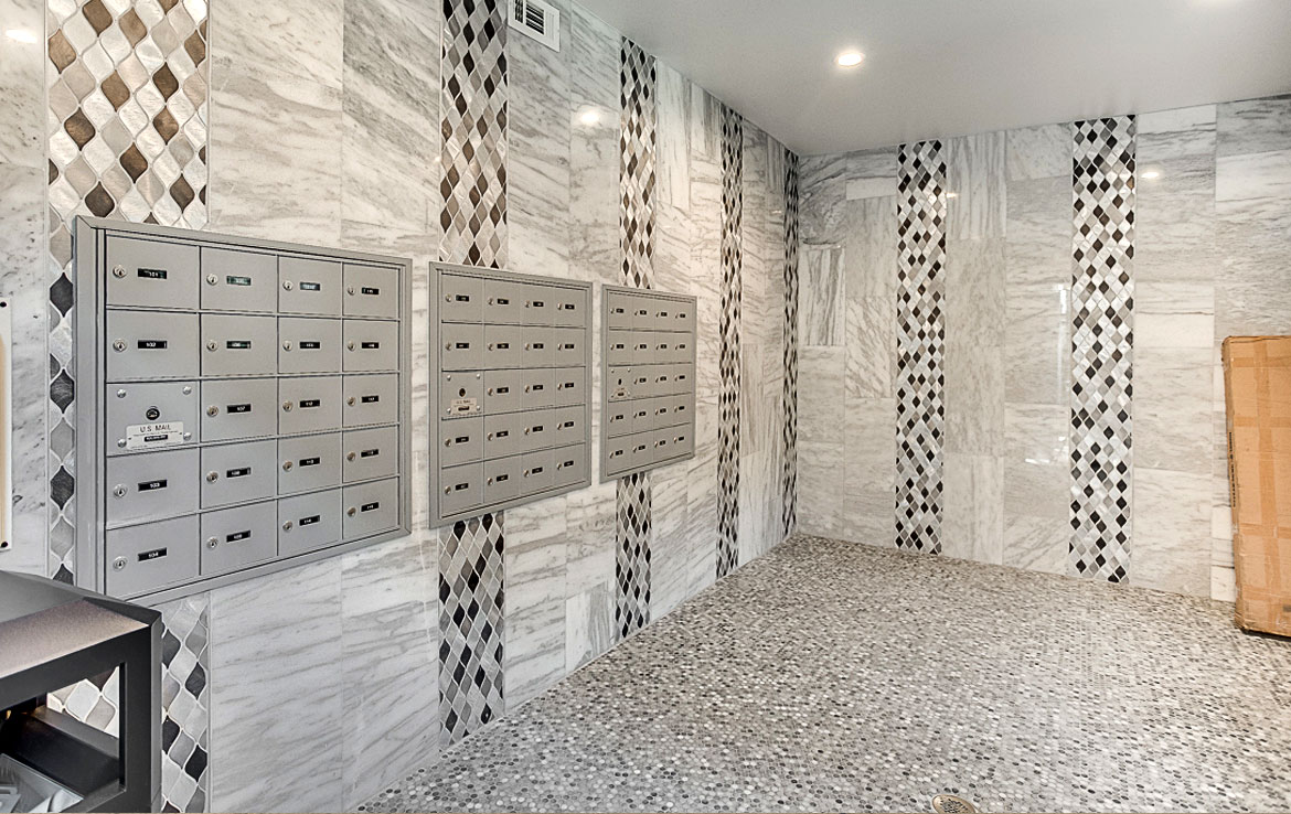 A well-lit room with walls and floors made of gray and white marble tiles, showcasing Orand Capital's exquisite attention to detail. Three sets of lockable mailboxes are mounted on the wall to the left. The decor includes vertical stripes of diamond-patterned tiles, reflecting our portfolio's elegance.