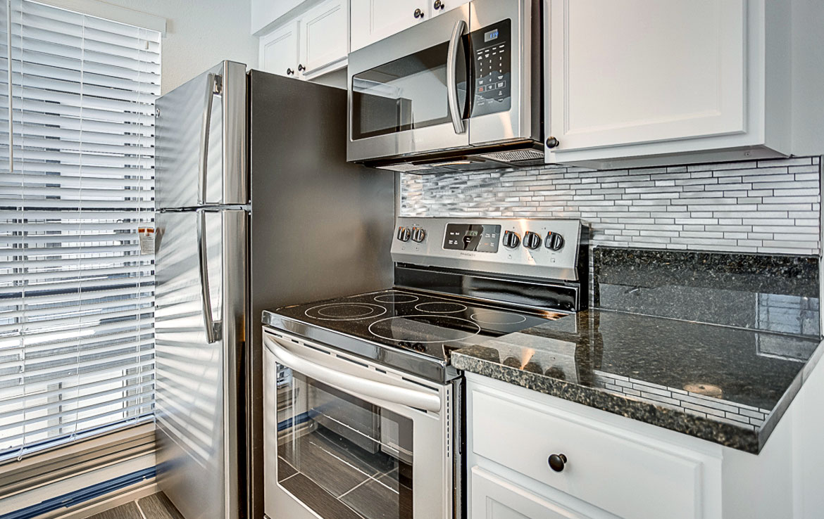 Modern kitchen with stainless steel appliances, including a refrigerator and range with a glass cooktop, beneath a built-in microwave. The white cabinets have dark countertops. A window with blinds is on the left side. The backsplash features small tiled patterns and is part of our portfolio at Orand Capital.