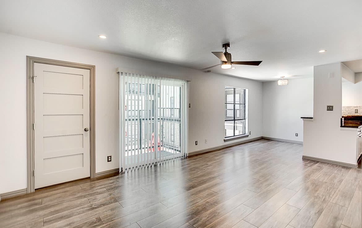 A modern, empty living room with white walls, wood flooring, and a ceiling fan. There's a large window with vertical blinds leading to a balcony on the left, a door beside it, and another large window near a partially visible kitchen on the right. Featured in Our Portfolio - Orand Capital.