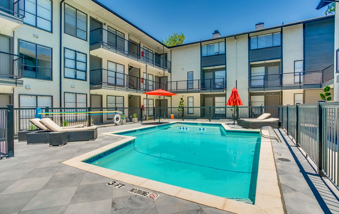 Orand Capital's portfolio includes a modern three-story apartment complex featuring a central outdoor pool surrounded by lounge chairs and umbrellas. The area is enclosed by a safety fence, and the building's balconies overlook the pool. The sky is clear, indicating a sunny day.