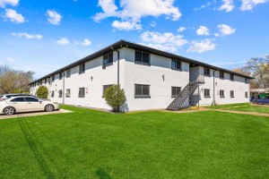 Our Portfolio features a two-story white apartment building with a staircase on the side, surrounded by a green lawn. The sky is clear with a few clouds, and a white car is parked next to the building on the left side, with trees visible in the background—Orand Capital.